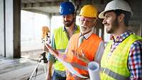 Drei Männer mit Warnwesten und Schutzhelmen blicken auf einer Baustelle auf einen Plan, im Hintergrund ein Vermessungsgerät