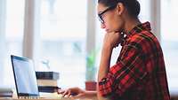 Dunkelhaarige Frau mit Brille und Flanellhemnd sitzt am Schreibtisch und arbeitet an einem Laptop